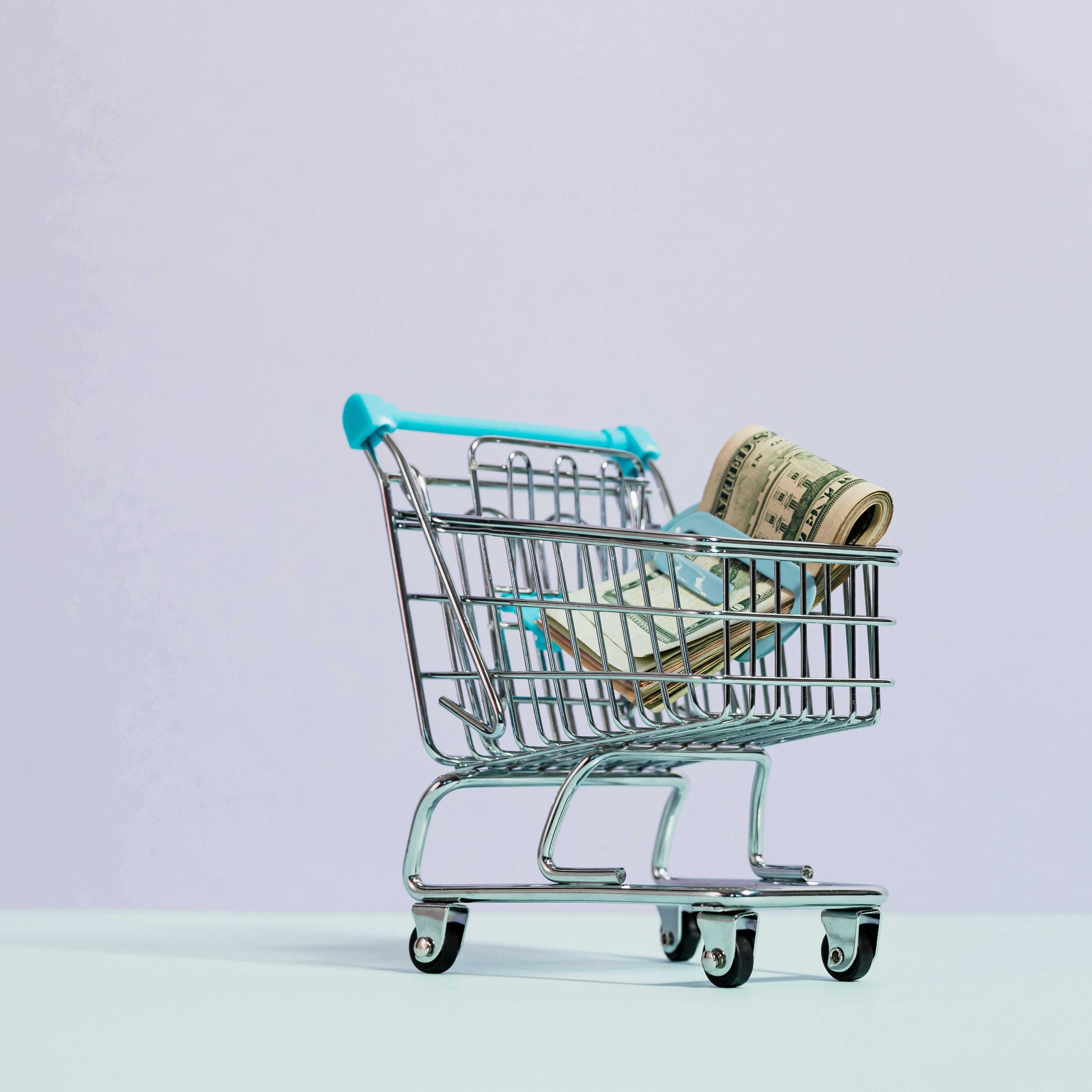 A small shopping cart filled with rolled-up paper currency is placed against a plain light purple background. The cart's handle is blue, and the light blue surface further enhances the minimalist aesthetic of the image.