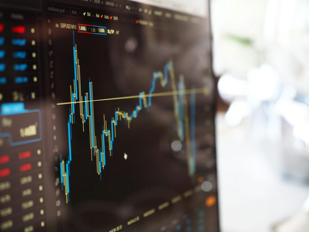 Close-up of a computer screen displaying financial data with a blue candlestick chart. There are various numerical values in red and green columns on the left side, while the chart depicts fluctuating market trends against a black background.