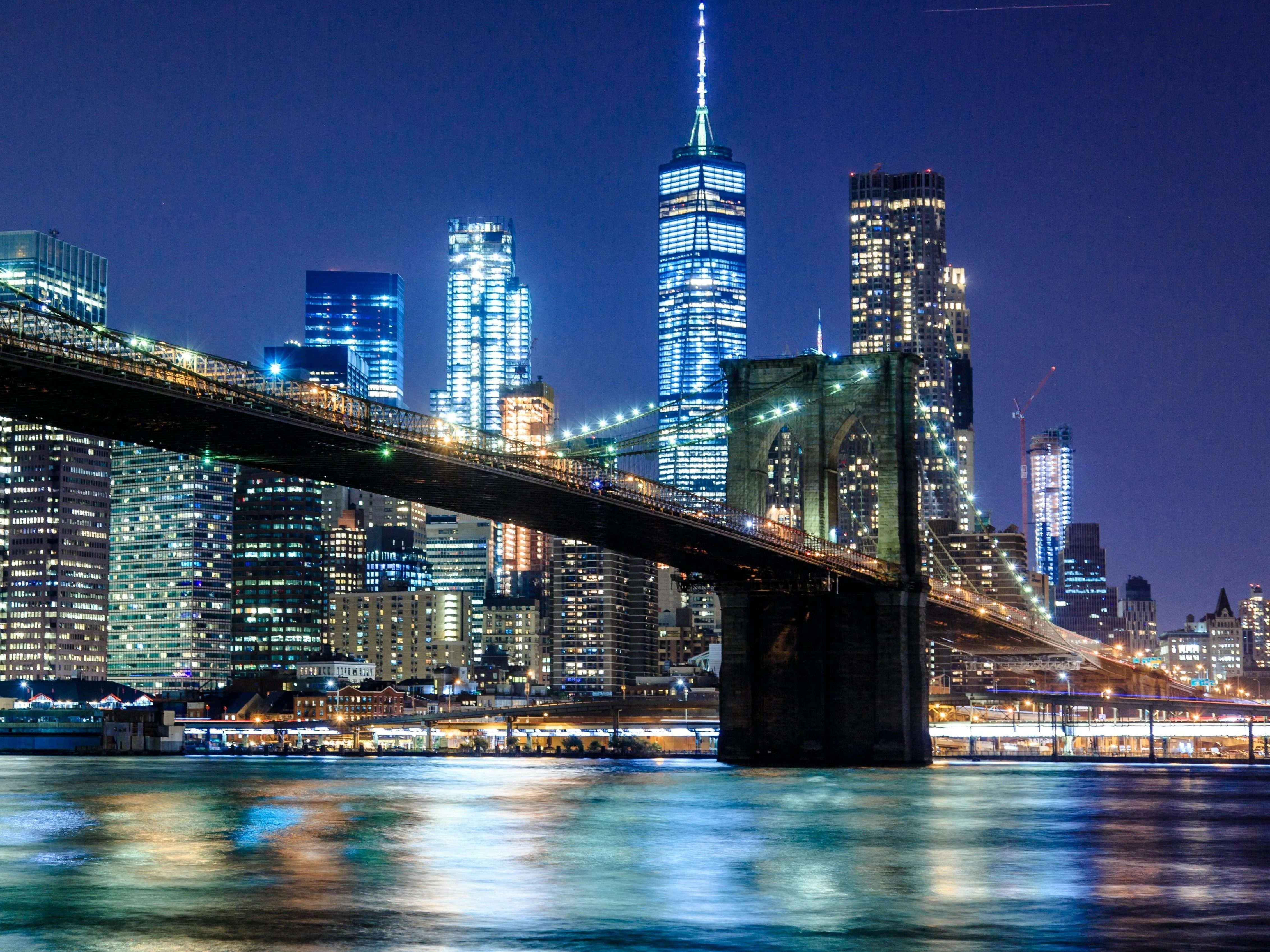 Night view of Brooklyn Bridge with illuminated skyscrapers of Manhattan in the background reflecting in the East River, New York City. The scene features a mix of modern and historic architecture under a clear night sky.