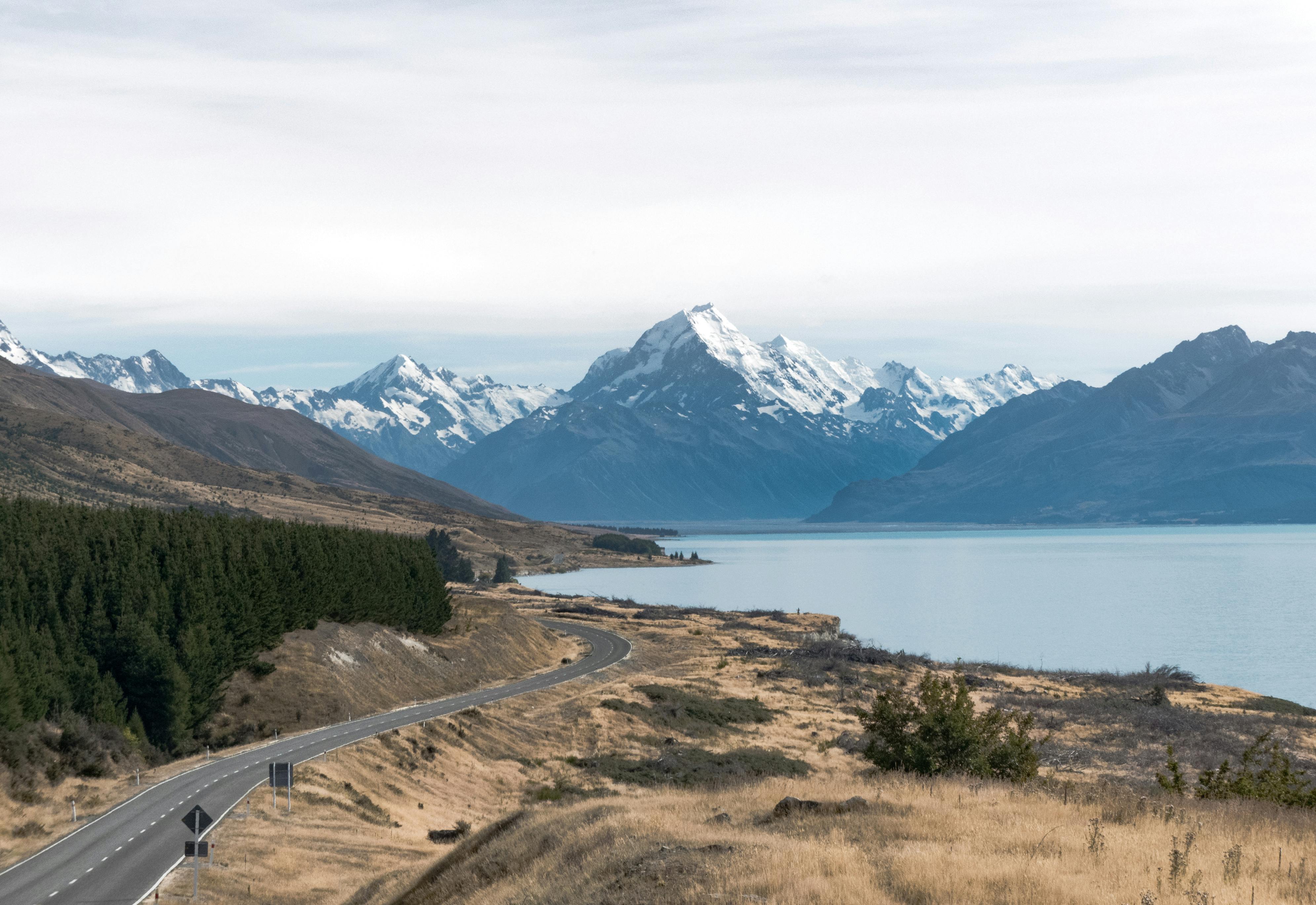 A scenic landscape featuring a winding road next to a large, calm blue lake. Tall, snow-capped mountains rise in the distance, and the surrounding terrain is a mix of grassy fields and dense patches of evergreen trees. The sky is mostly clear with a few wispy clouds.