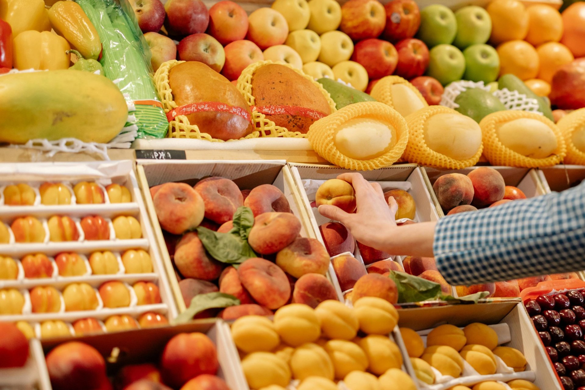A person’s hand picks up a peach from a display of neatly arranged fruits at a market. The display includes various types of peaches, apples, papayas, and other colorful fruits. The produce is organized in wooden crates and baskets.
