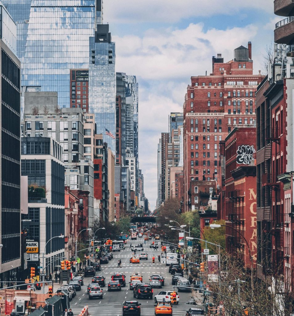 A bustling city street lined with tall buildings, both modern glass skyscrapers and older brick structures. The street is busy with cars and taxis, and there are traffic lights and pedestrians along the sidewalks. The sky is partly cloudy.