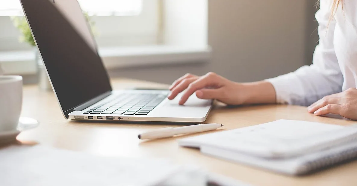 A person wearing a white shirt is seated at a wooden table, typing on a laptop. The table has notes and a pen. Sunlight comes through a window in the background, providing natural light to the scene.