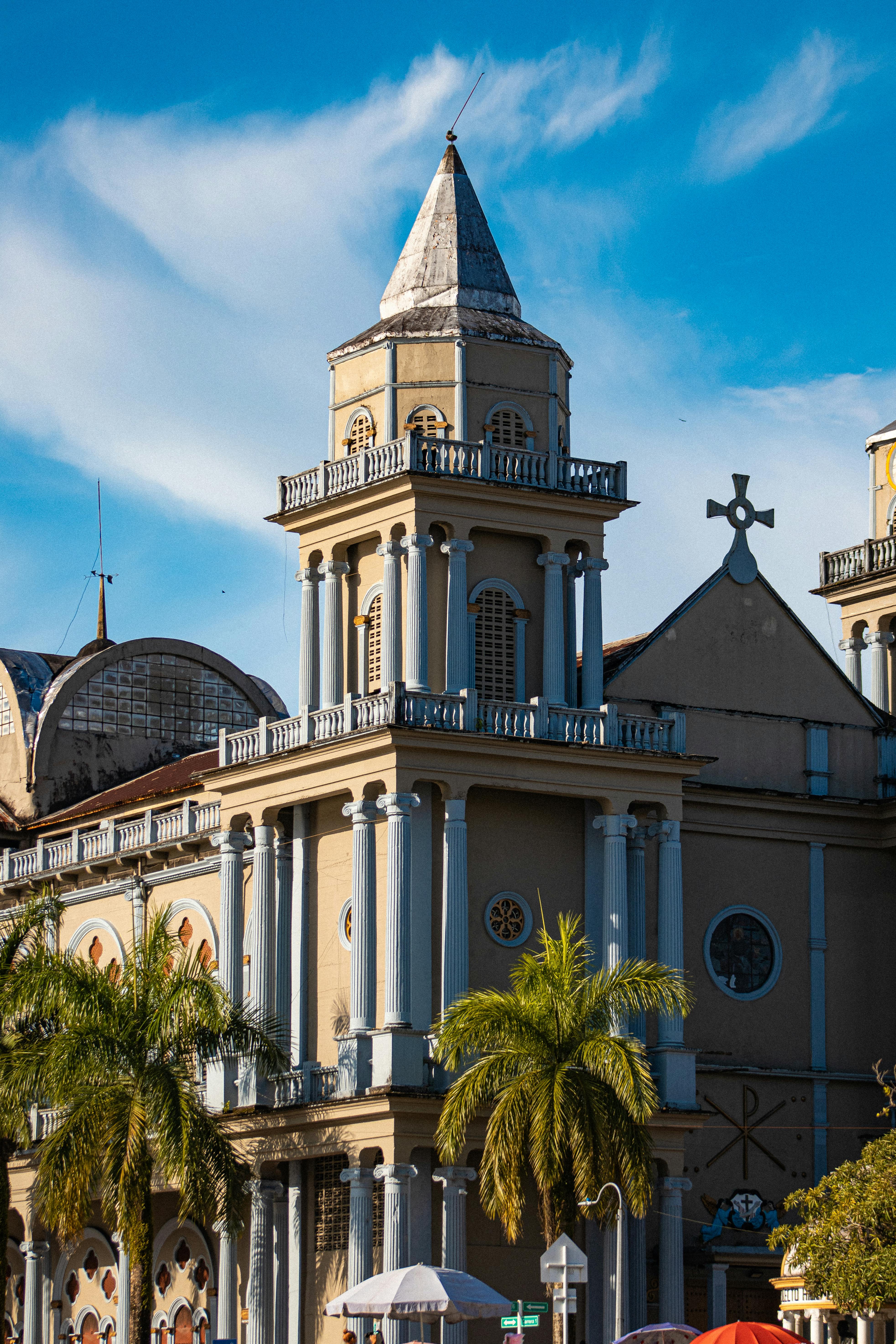 A beige-colored church with a tall, pointed bell tower stands under a clear blue sky. The facade features circular windows and columns. Palm trees are in the foreground, adding a tropical touch to the scene.