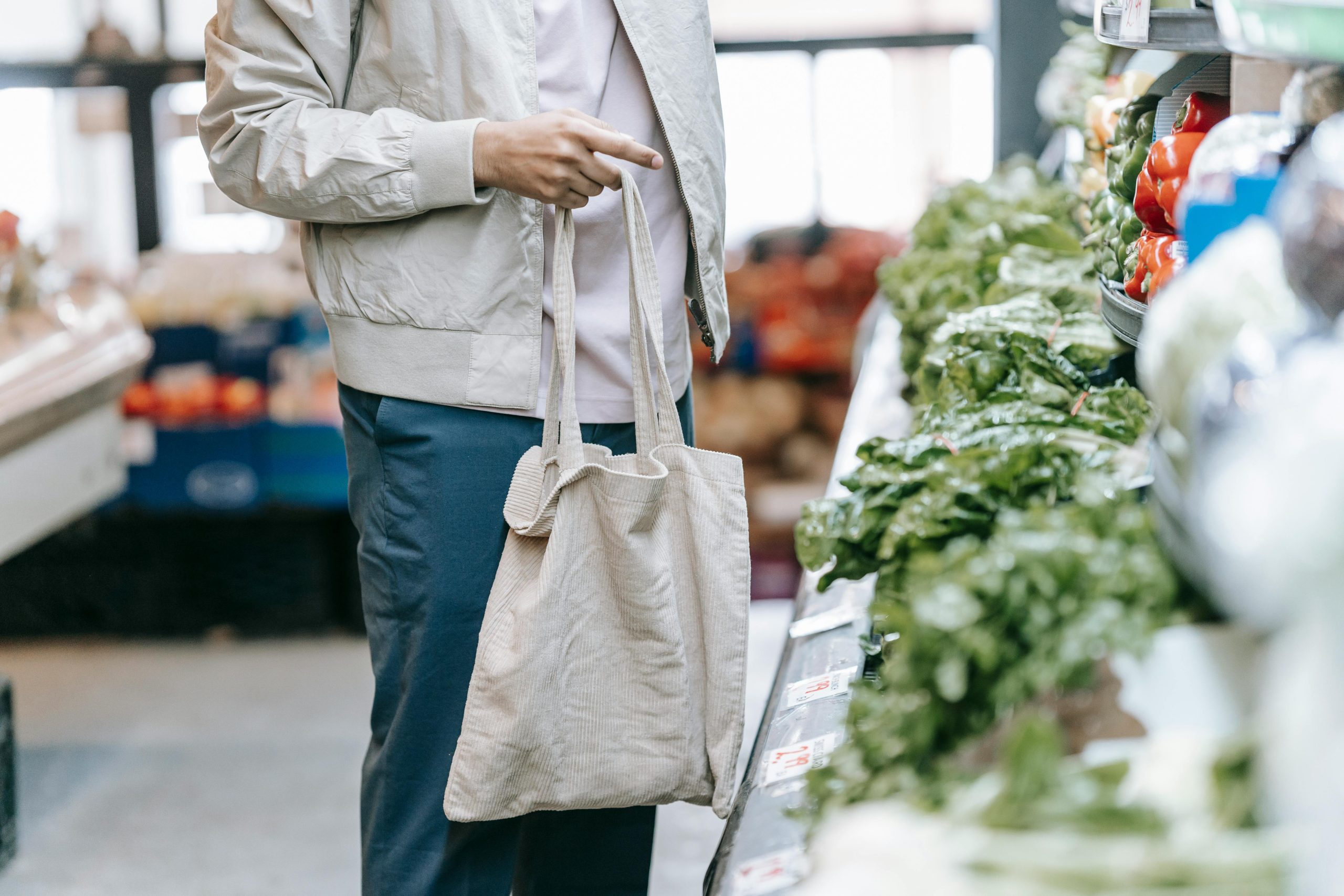A person wearing a beige jacket and holding a reusable tote bag is shopping for fresh produce in a grocery store. They are standing in front of a display of green leafy vegetables and bell peppers.