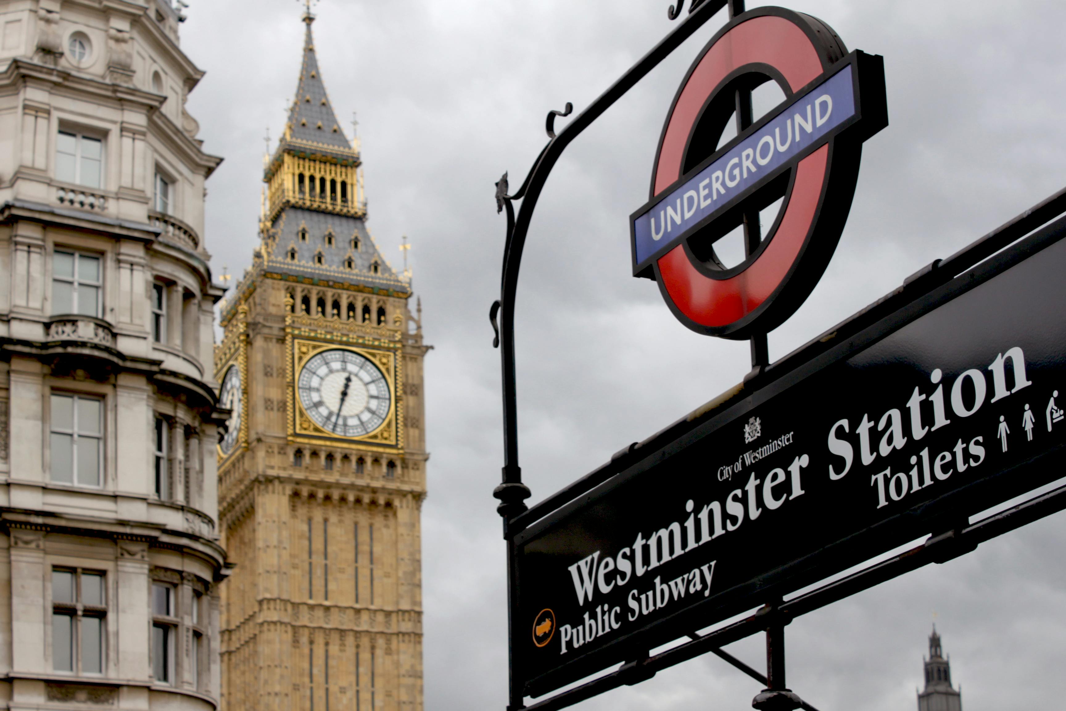 Image features the iconic Big Ben clock tower in London, with the Westminster Station Underground sign prominently displayed in the foreground. The sky is overcast, and in the background, a historic building is partially visible next to Big Ben.